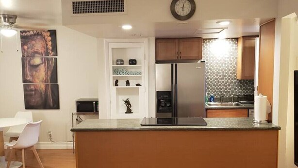 A modern kitchen with a central island, wooden cabinets, a stainless steel refrigerator, and a wall-mounted clock.