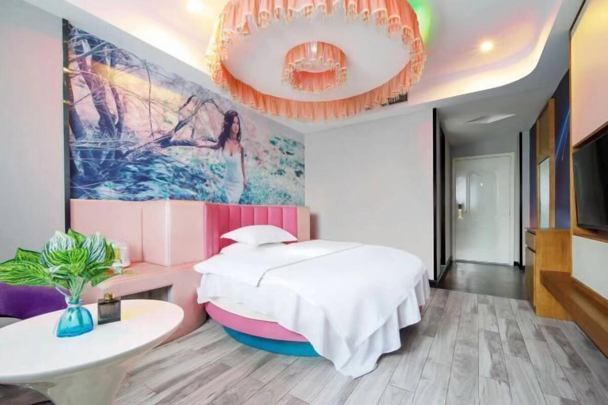 A modern bedroom with a bed, a round table with a plant, a large painting, and a unique ceiling light fixture.
