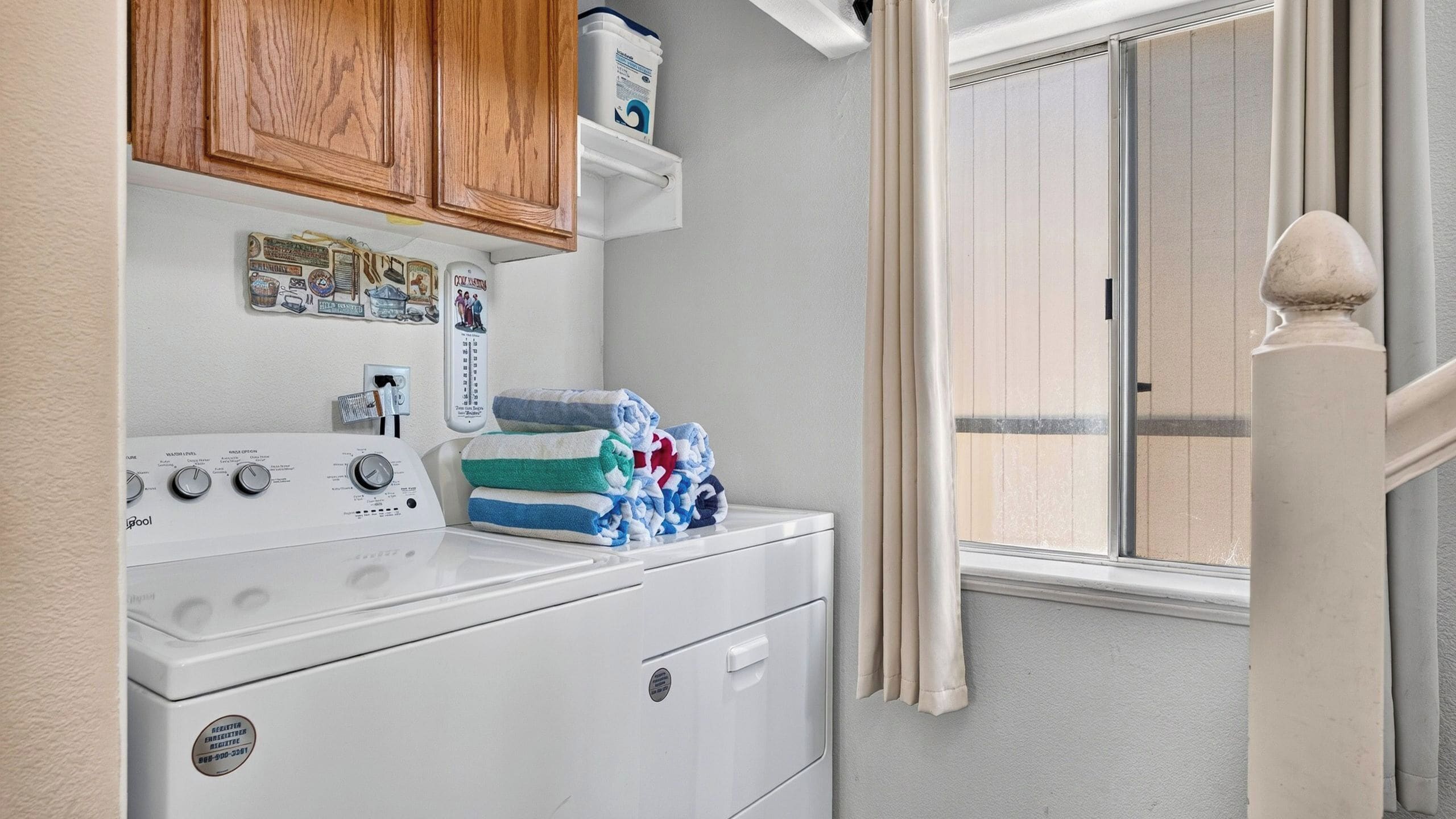 Un espace buanderie équipé d’un lave-linge et d’un sèche-linge à chargement frontal, de meubles en bois, d’un thermostat et d’une fenêtre avec des stores.