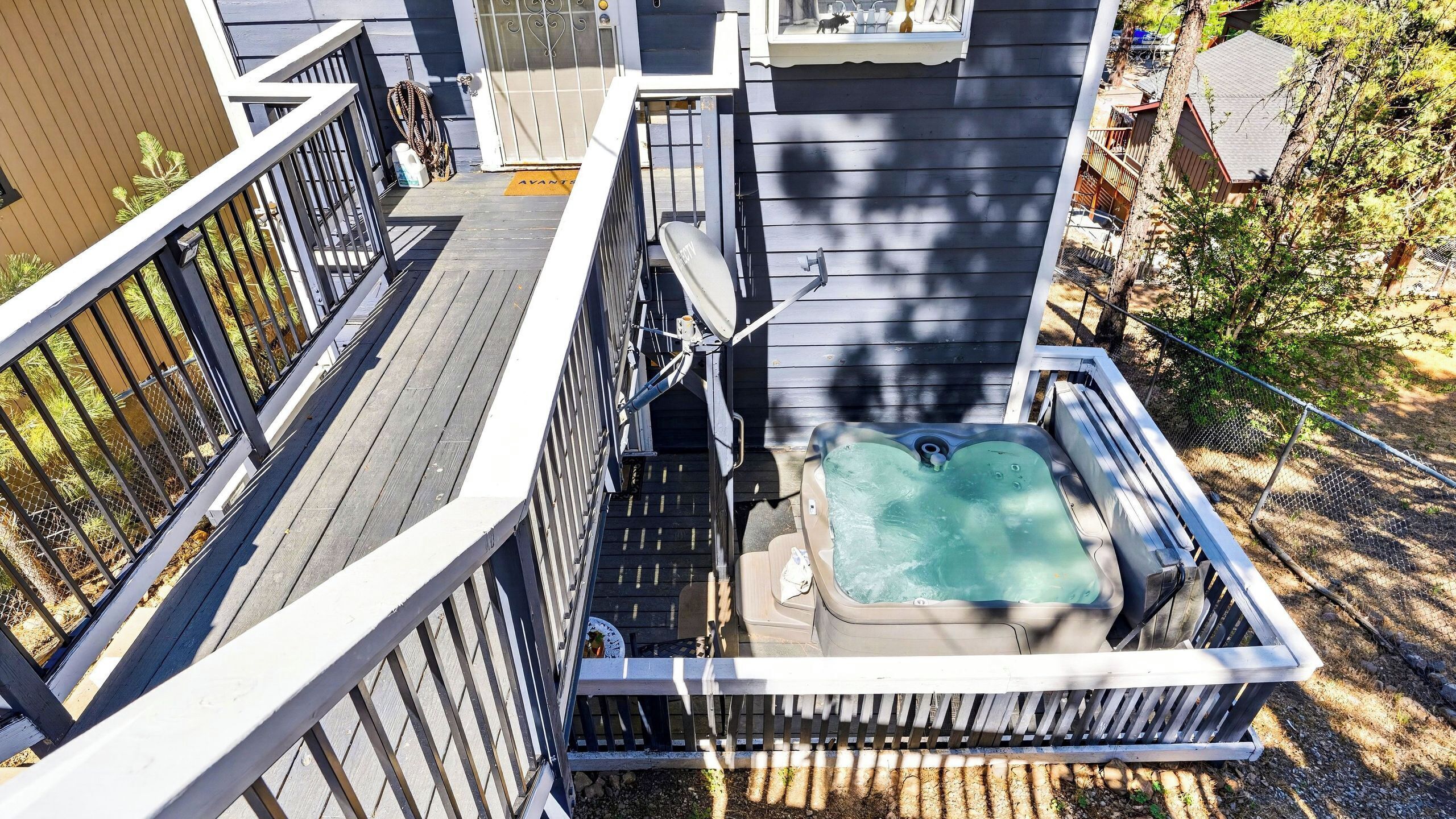 A hot tub on a wooden deck with a metal railing, surrounded by trees and a fence.