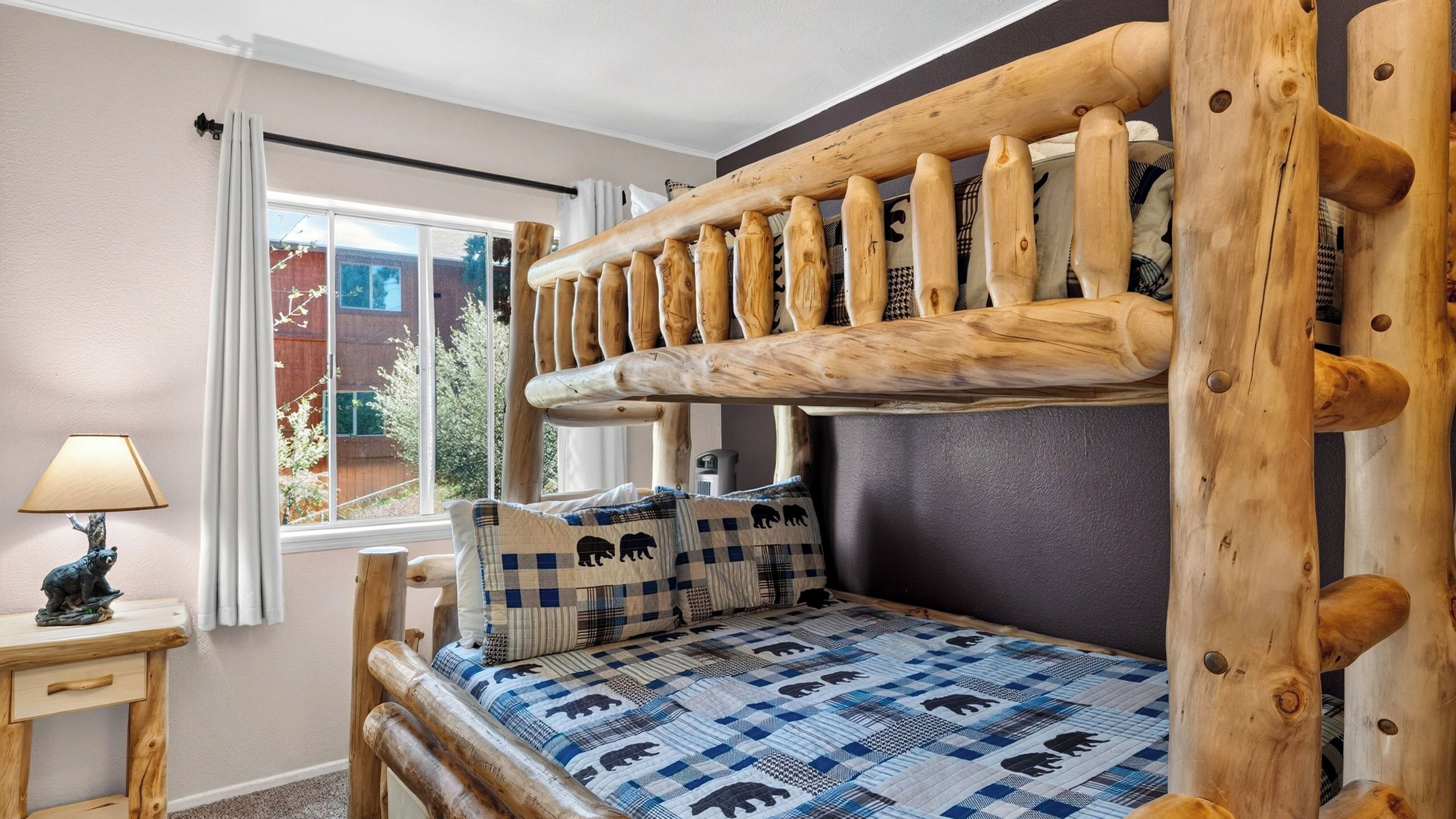 Ein Stockbett mit Holz Kopfteil, ein Fenster mit Vorhängen, ein Nachttisch mit Lampe und eine gemusterte Bettdecke.