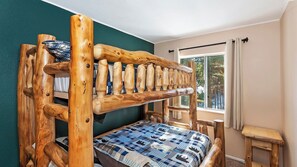A bunk bed with a checkered bedspread, a small wooden stool, and a window with curtains.