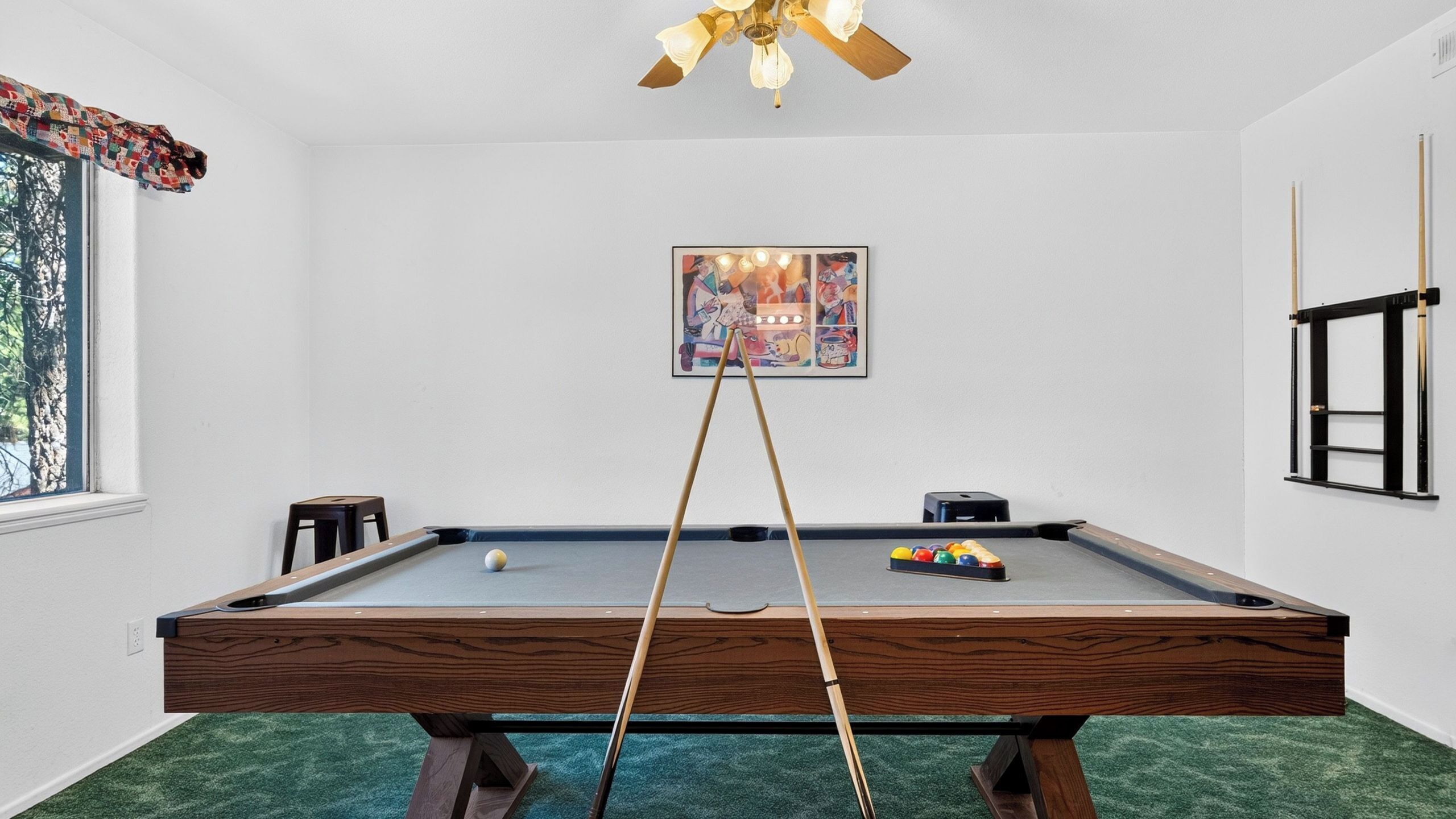 A pool table with a cue stick, a framed picture, and a window with a view of trees.