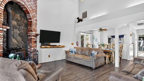 A living room with a stone fireplace, a flat-screen TV, a sofa, and a dining area. - Mountain Palette by Avantstay Hot Tub, Game Room, Deck, Sweeping Views! (Big Bear City)