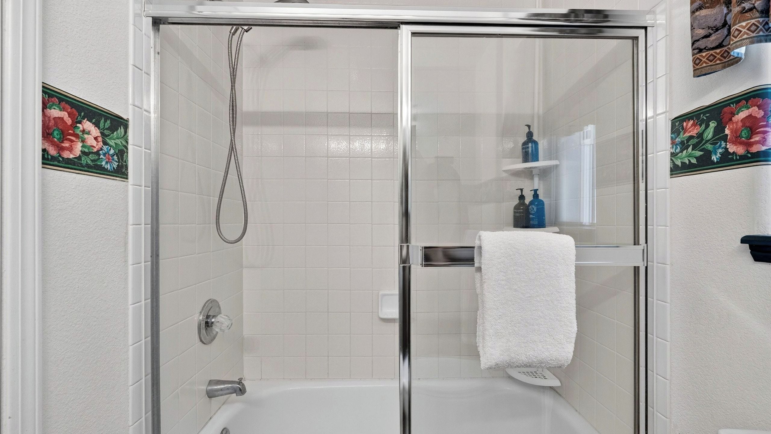 Douchecabine met glazen wand, een handdouche, een ligbad en een witte handdoek aan een rek.