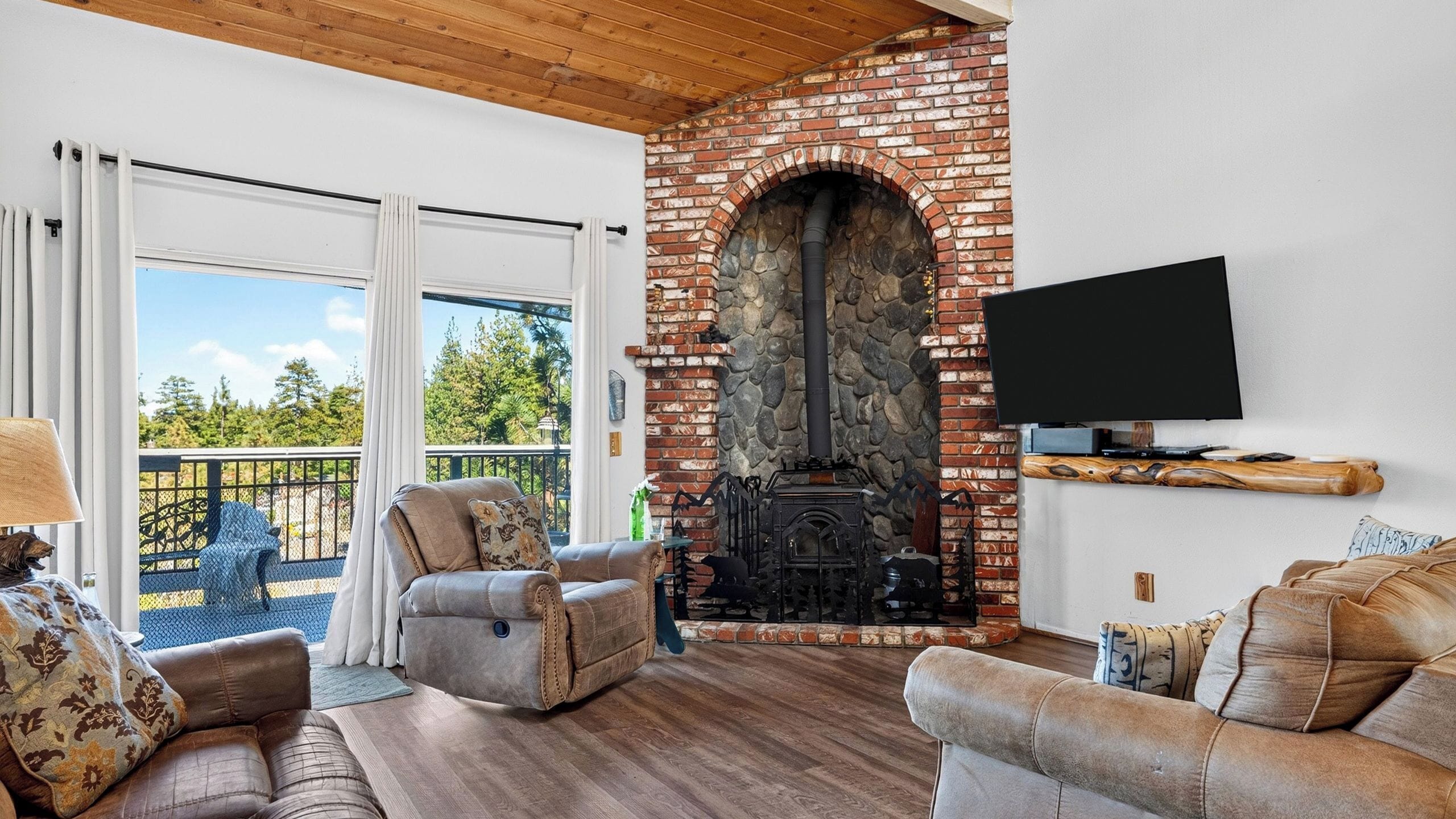 A living room with a stone fireplace, a TV mounted on the wall, and a balcony with outdoor furniture.