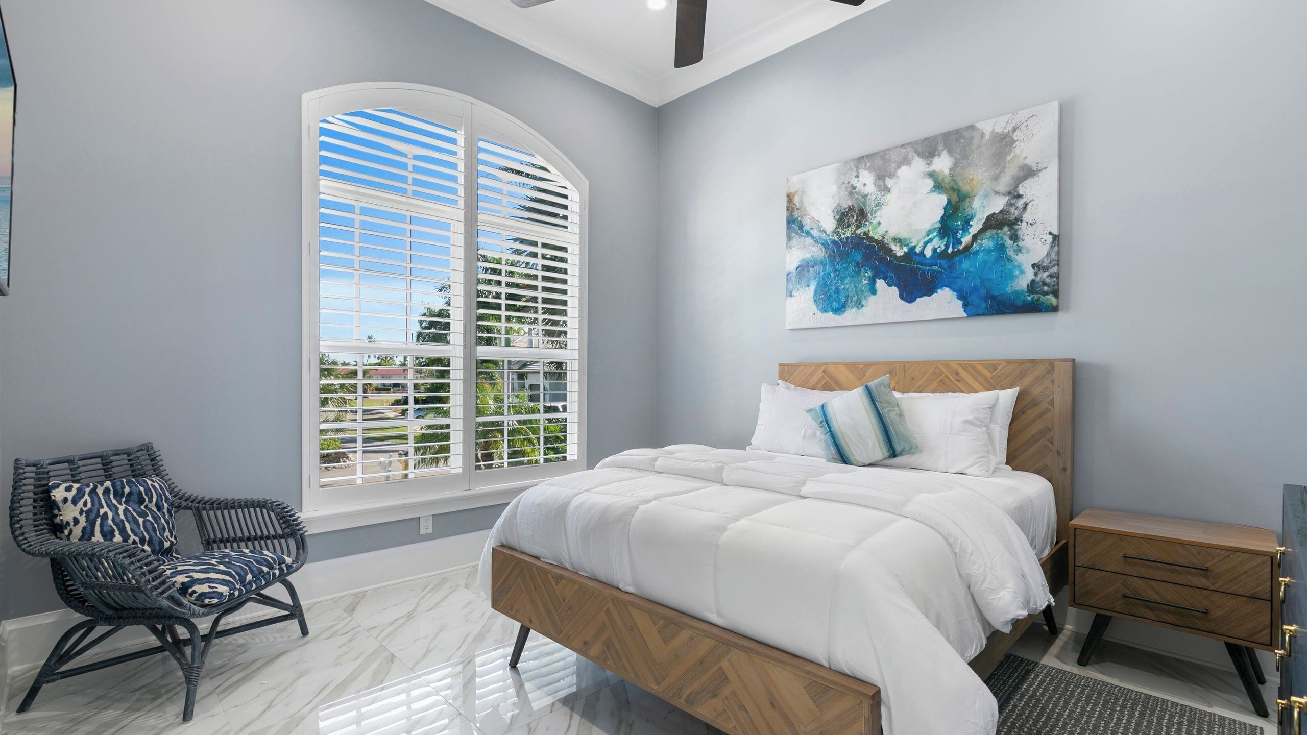 A bedroom with a bed, a chair, a nightstand, a painting, and a window with shutters.