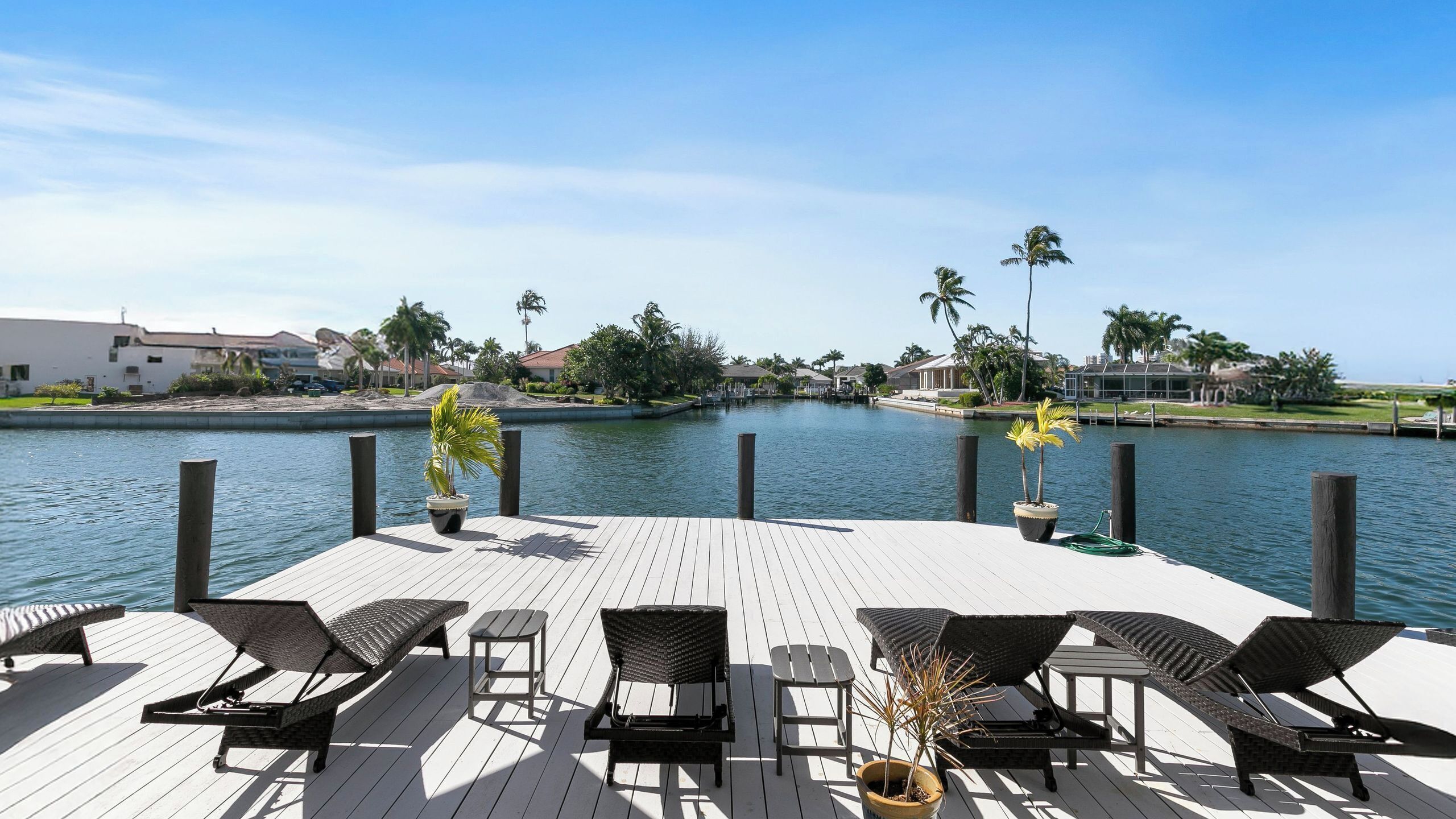 A wooden deck with lounge chairs and potted plants overlooking a calm water body with palm trees and houses in the background.