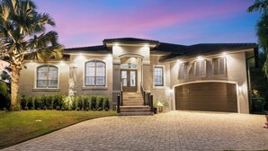 A large, two-story house with a driveway leading to a front door, surrounded by a well-maintained lawn and palm trees.