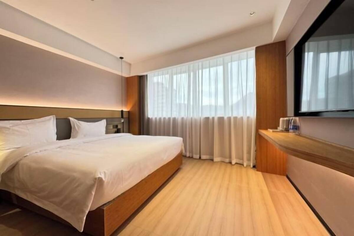 Quarto de hotel moderno com uma cama grande, piso de madeira, escrivaninha embutida e uma janela ampla com cortinas.