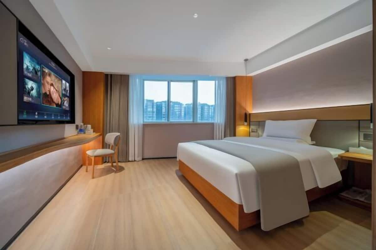 Quarto de hotel com uma cama grande, televisão presa na parede, janela com vista para a cidade e um pequeno espaço para sentar.