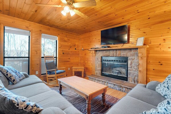 A cozy cabin interior with a stone fireplace, wooden walls, a television mounted above it, and a comfortable seating area with a sofa and a rocking chair.
