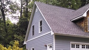 A two-story house with a grey roof and white trim, surrounded by lush greenery and flowering plants. - Great Asheville Location (Asheville)