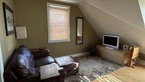 A cozy loft bedroom with a sloped ceiling, a large window with blinds, a flat-screen TV, a wooden dresser, a leather sofa, and a small table. - Great Asheville Location (Asheville)