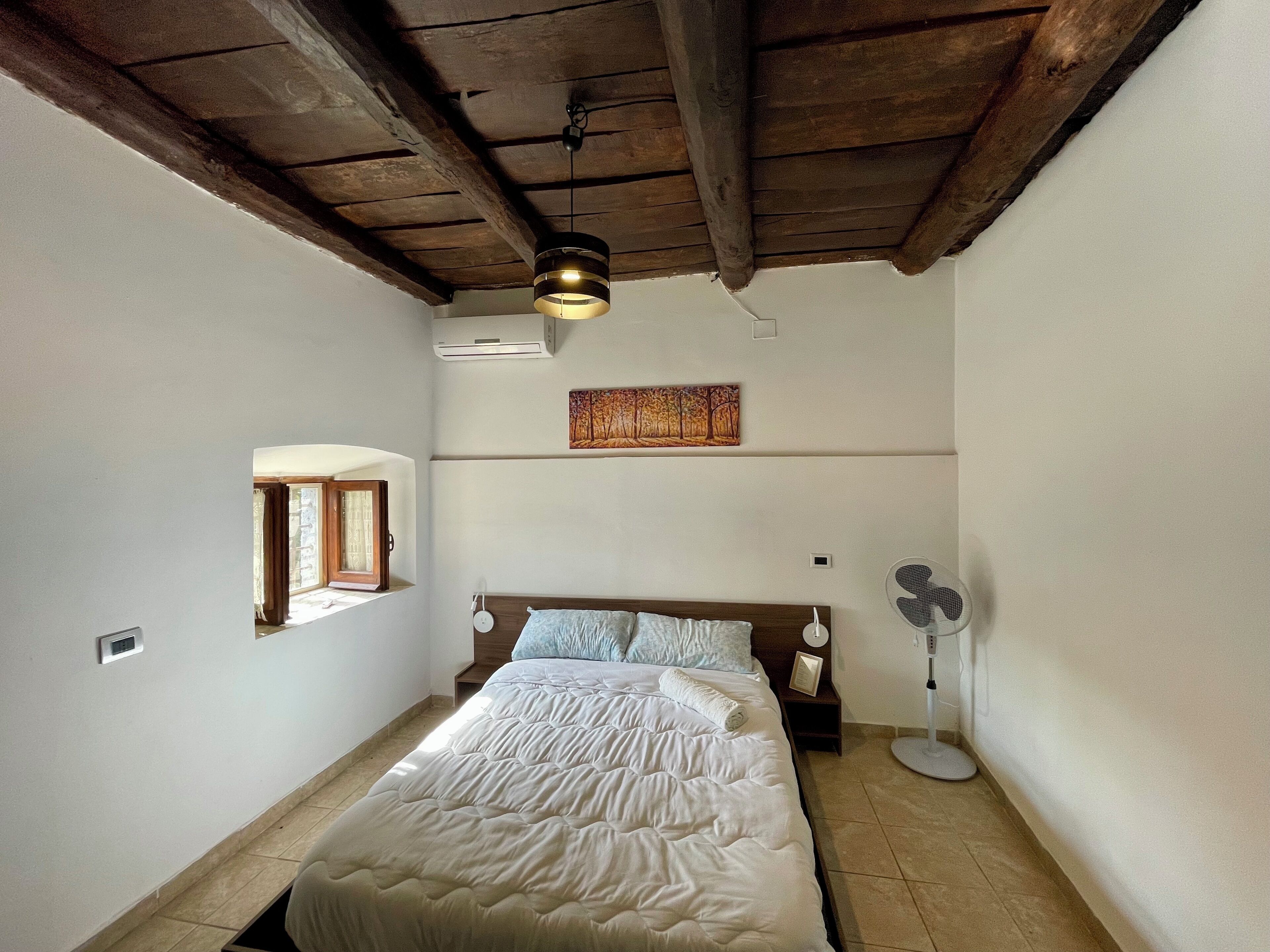 A bedroom with a bed, a ceiling fan, an air conditioning unit, a window, and a painting on the wall.