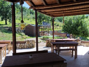 Un patio al aire libre con mesas y bancos de madera, un muro de piedra y vistas a un edificio rústico y follaje en el fondo.