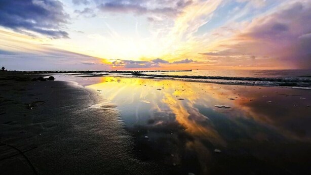 Una playa con un atardecer, aguas tranquilas y un muelle a lo lejos.