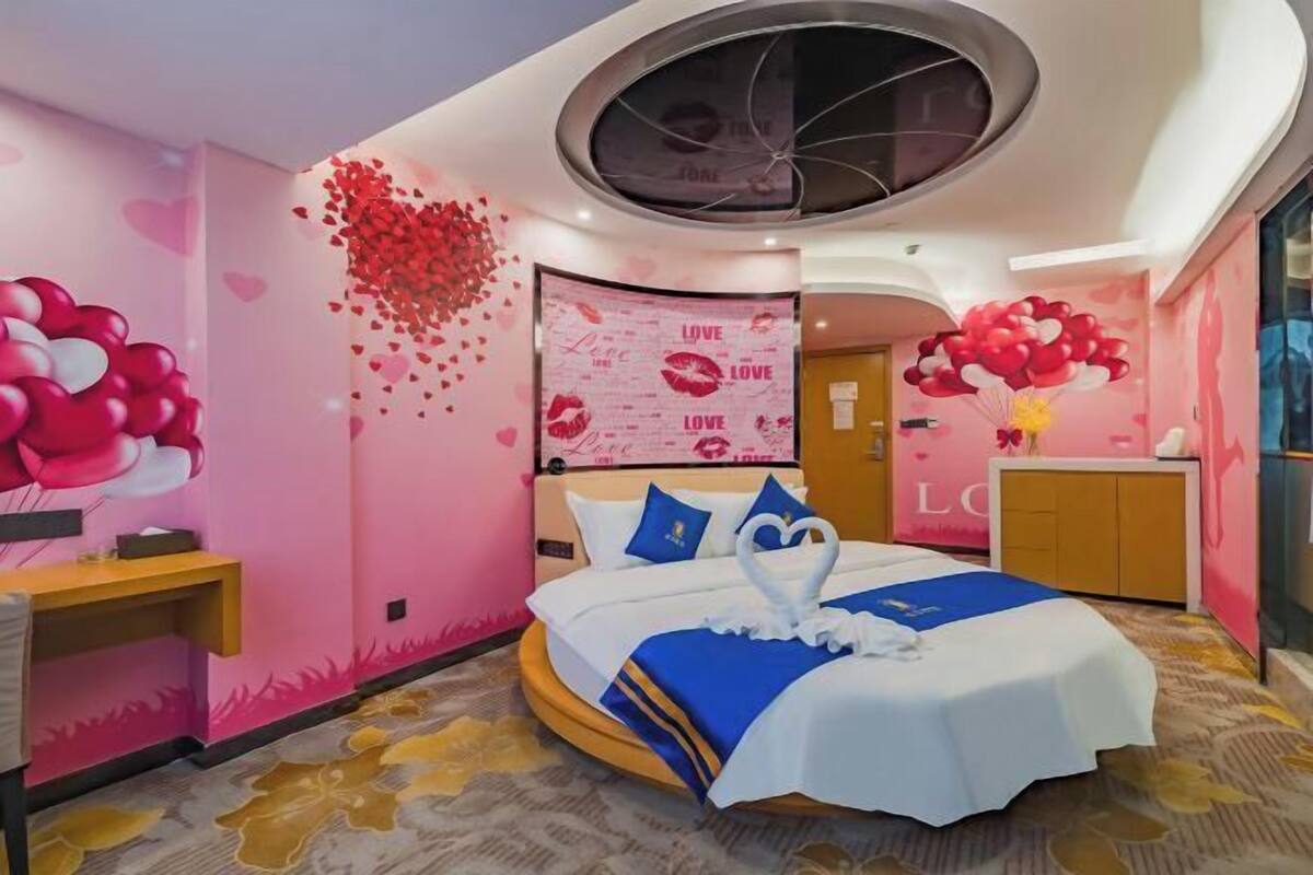 A pink-themed hotel room with a bed, a desk, and a wall decorated with hearts and the word "LOVE".