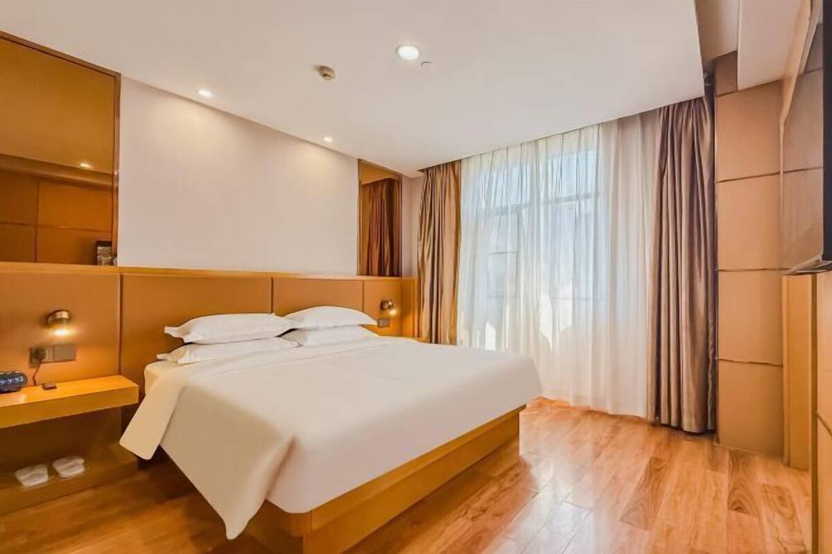 A modern hotel room with a wooden bed, bedside table, and a large window with curtains.