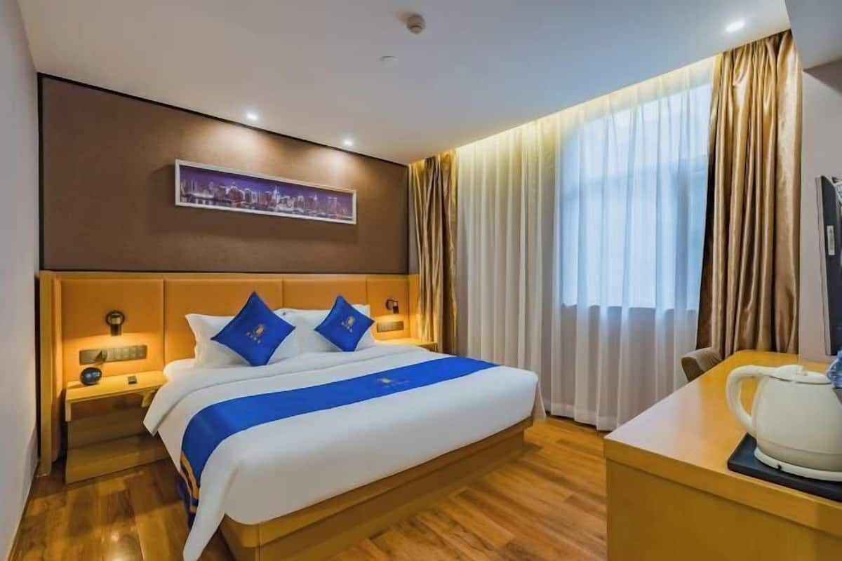 A hotel room with a bed, bedside tables, a desk with a kettle, a painting on the wall, and curtains.