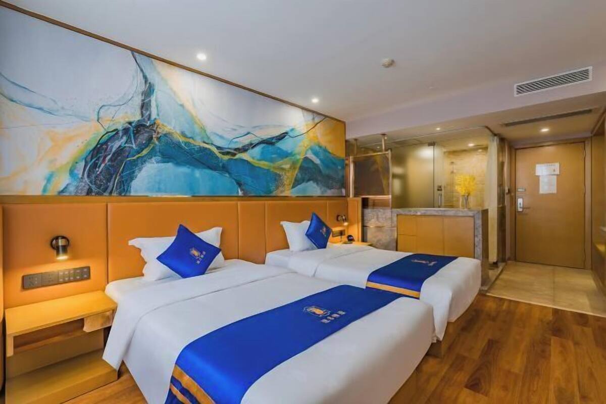 A hotel room with two beds, a large abstract painting on the wall, a minibar, and a bathroom visible through an open door.