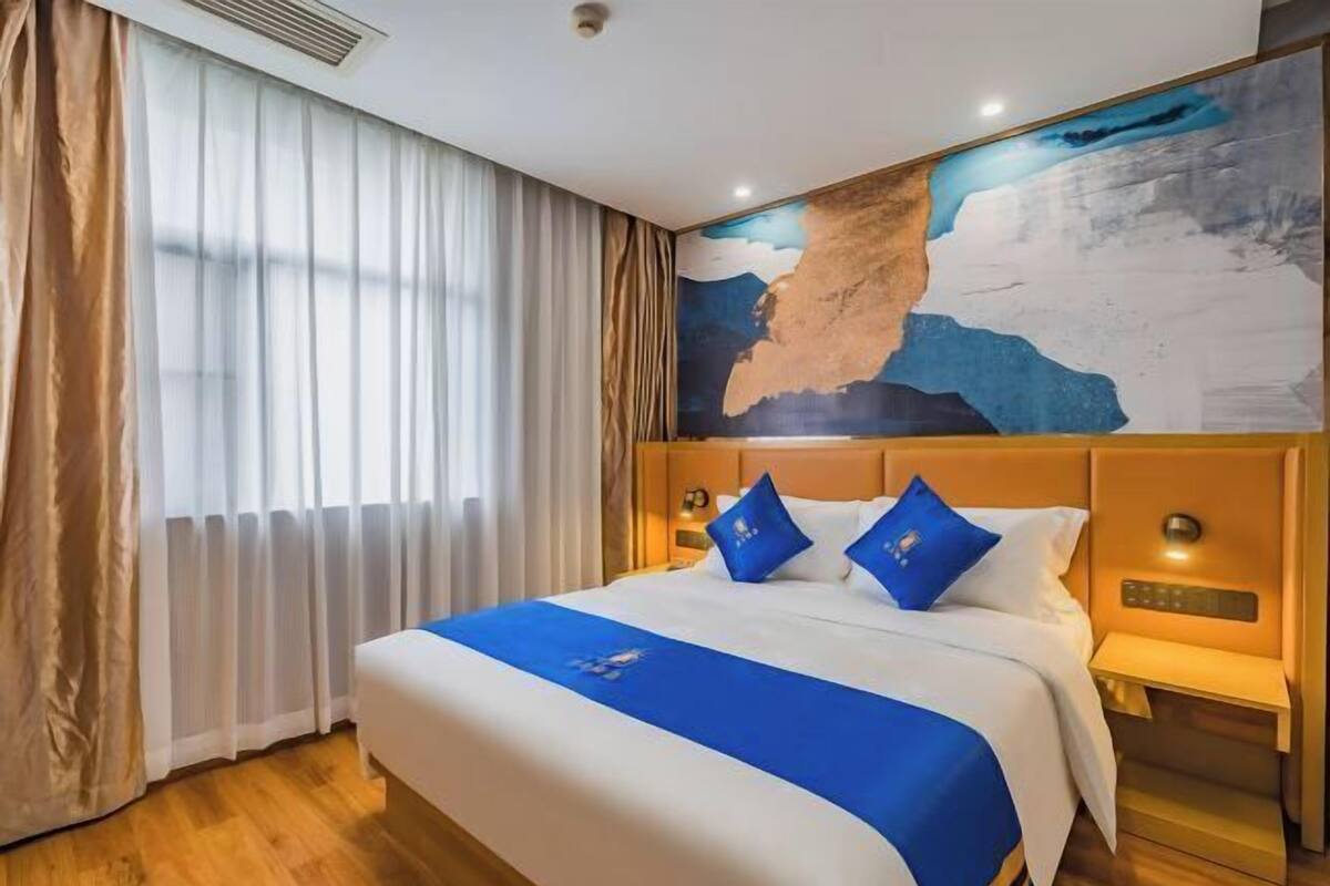 A hotel room with a large bed, a window with sheer curtains, a wall painting, and a nightstand with a lamp.