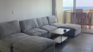 Un salon moderne avec un canapé d’angle gris, une petite table basse et un ventilateur de plafond. Une porte-fenêtre coulissante donne accès à un balcon où se trouvent des chaises en bois.