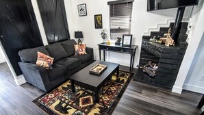 Un salon moderne avec un canapé de couleur sombre, une table basse et un tapis à motifs. On y trouve un escalier orné d’une pancarte « Bienvenue » et d’une petite statue.