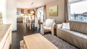 A modern living room with a sofa, a dining table with chairs, and a kitchen area in the background. - Maeterlinck 201 Sunny Penthouse in Koksijde (Koksijde)