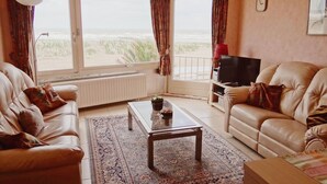 Una sala de estar con dos sofás de cuero, una mesa de centro, un televisor sobre un mueble bajo y un ventanal con cortinas.
