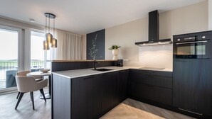 A modern kitchen with dark cabinetry, a central island, a built-in oven, and a dining area with chairs and a table. - Loft Apartment by Veerse Meer Marina (Kamperland)