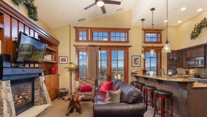 A living room with a fireplace, a television, a sofa, and a kitchen area with a bar. - Ultimate ski-in, ski-out!  Steps from ski lifts & gondola. Resort amenities! Air Conditioning! (Breckenridge)