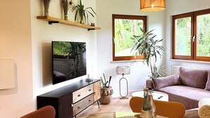 Un soggiorno moderno con un tavolo da pranzo in vetro, un divano, una televisione e un'ampia finestra vista verde.