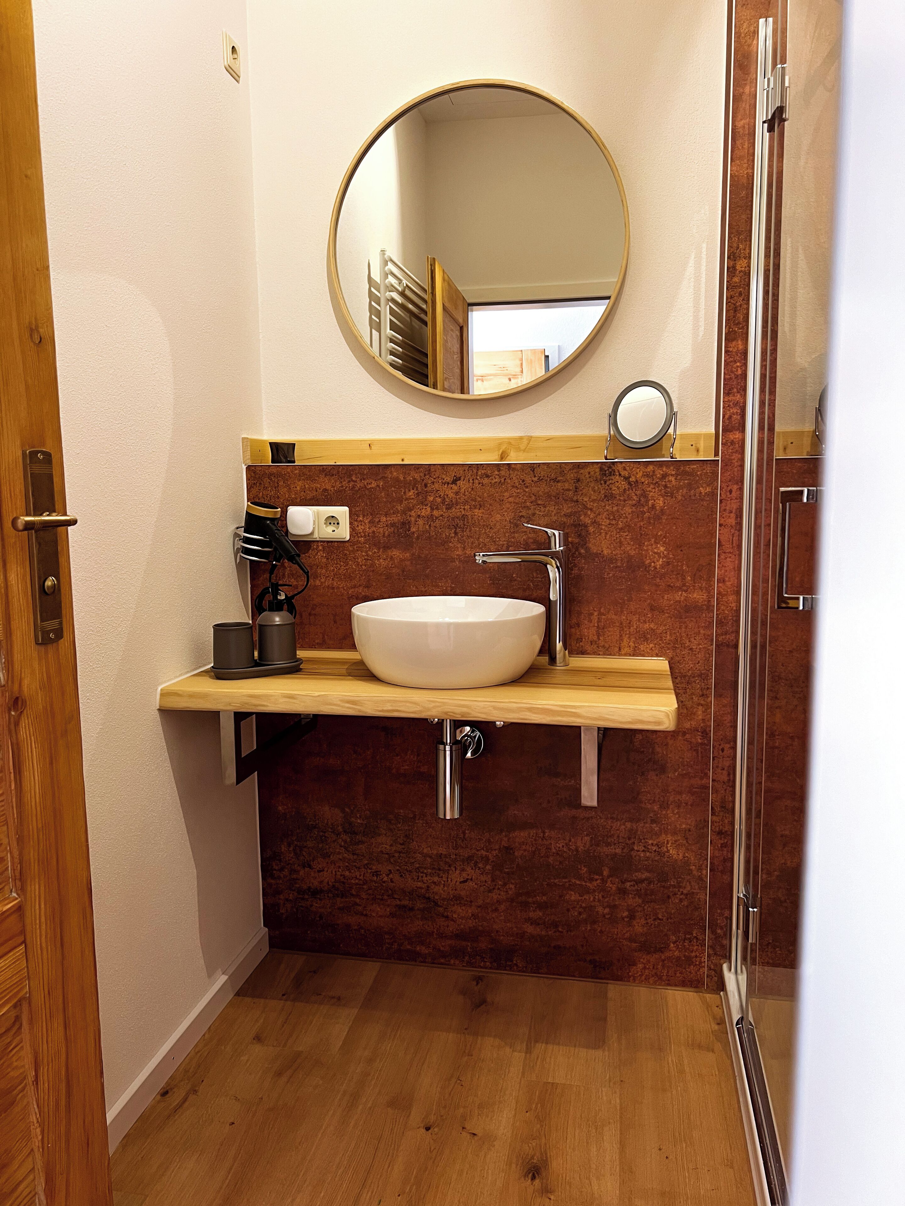 A bathroom with a wooden vanity, a white sink, a mirror, and a shower area.
