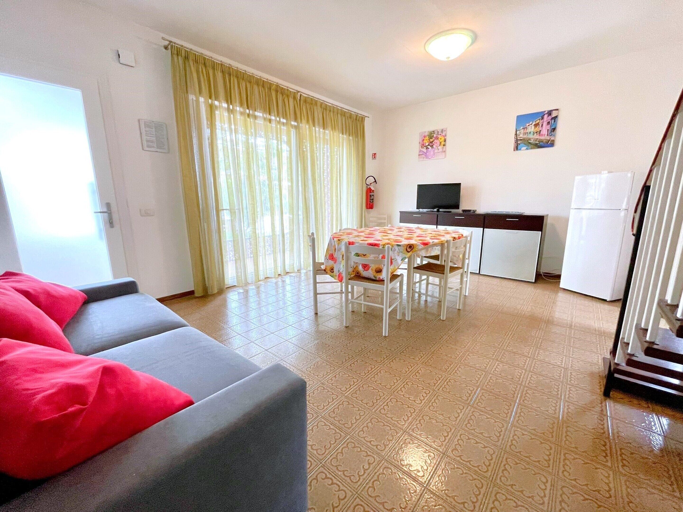 A compact living space with a sofa, a dining table with chairs, a kitchen area with a refrigerator, and a staircase.