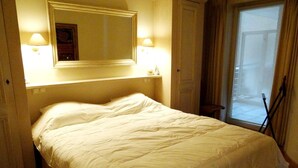 A neatly made bed in a room with a large mirror above it, a small shelf, and a window with curtains. - Seaside Comfort With Parking (Nieuwpoort)