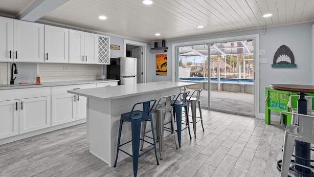 A modern kitchen with white cabinets, a large island with bar stools, and a view of a pool through glass doors. - HEATED POOL screened in - PET - TIKI - 4 Kayaks - Private Fishing & Boating Dock (Hernando Beach)