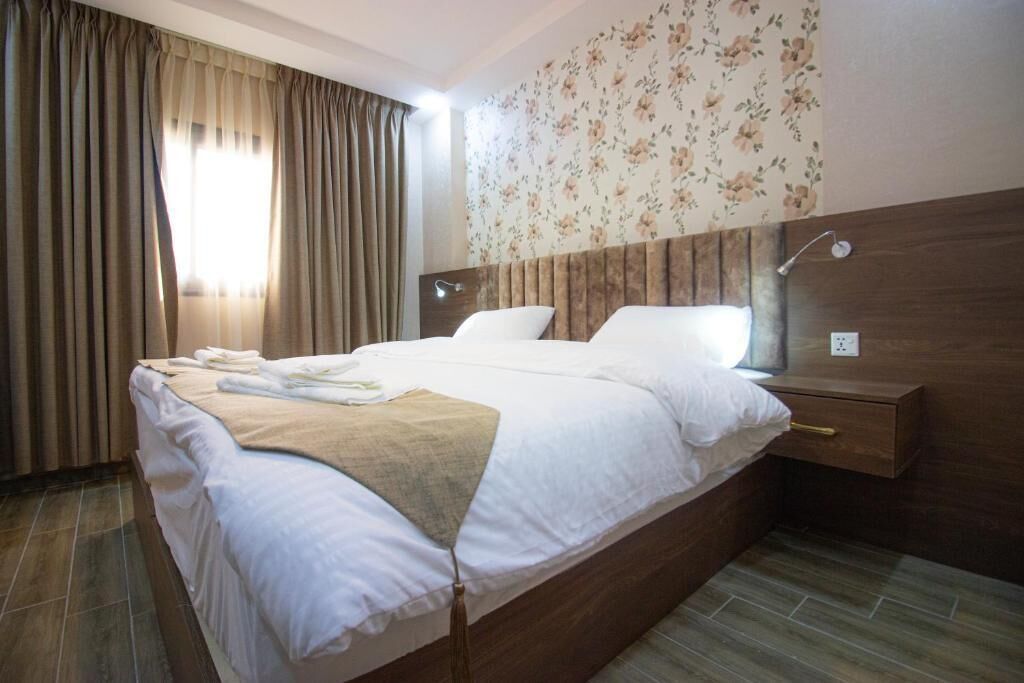 A neatly made bed with white linens and a beige throw blanket, a wooden headboard, a bedside table with a lamp, and a floral wallpaper pattern on the wall.
