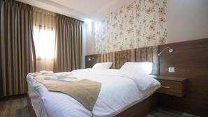 A neatly made bed with white linens and a beige throw blanket, a wooden headboard, a bedside table with a lamp, and a floral wallpaper pattern on the wall. - Beirut Hotel Two (Amman)
