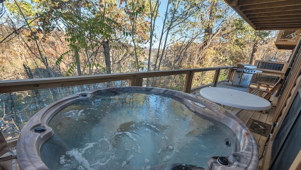 A hot tub on a wooden deck with a view of trees and a grill area.