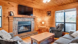 A cozy cabin interior with a stone fireplace, a flat-screen TV mounted above it, a comfortable gray sofa set, and a ceiling fan.