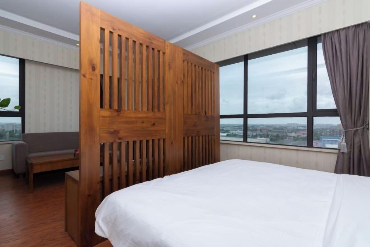 A hotel room with a bed, a wooden partition, a window with a city view, and a sofa.