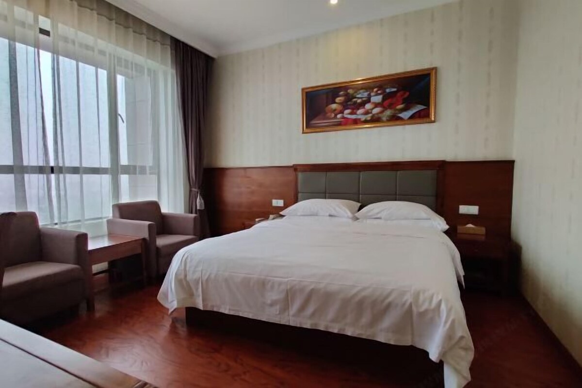 A hotel room with a bed, two chairs, a table, a painting on the wall, and a window with curtains.
