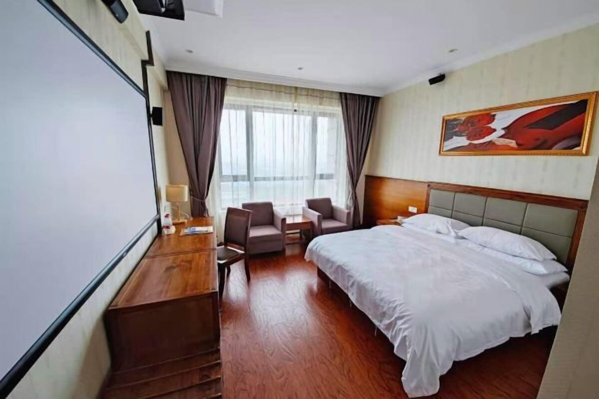 A hotel room with a bed, a desk, a chair, a window with curtains, and a painting on the wall.