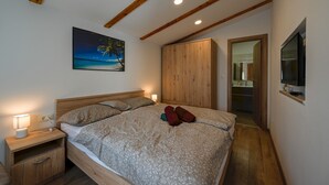 A bedroom with a wooden bed, a nightstand, a wooden wardrobe, a painting of a beach, and a TV mounted on the wall.