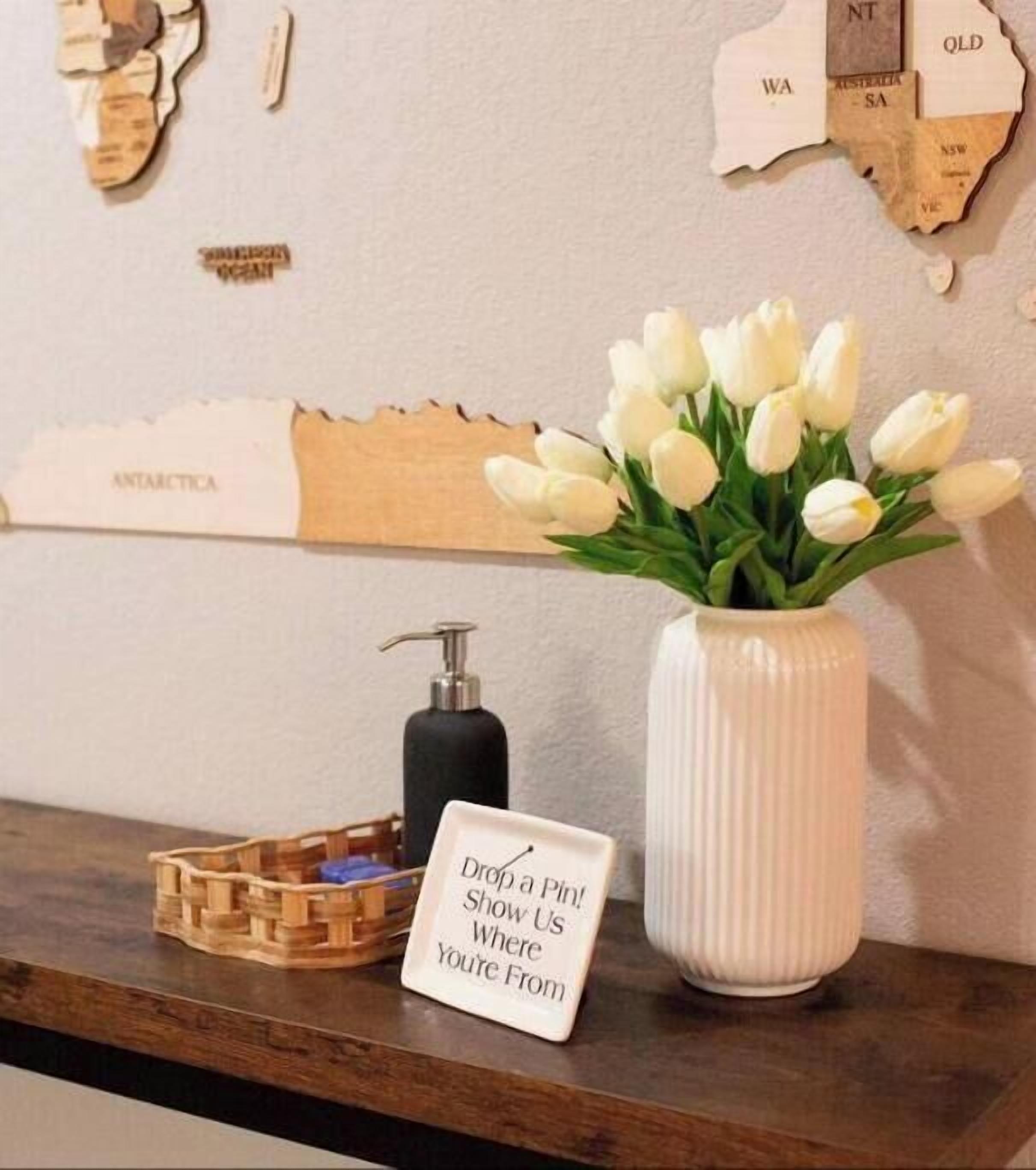 A wooden wall map with state names, a white vase with white flowers, a soap dispenser, and a sign that says "Drop a Pin! Show Us Where You're From."