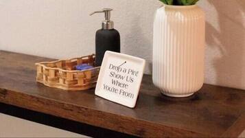 A wooden wall map with state names, a white vase with white flowers, a soap dispenser, and a sign that says "Drop a Pin! Show Us Where You're From."