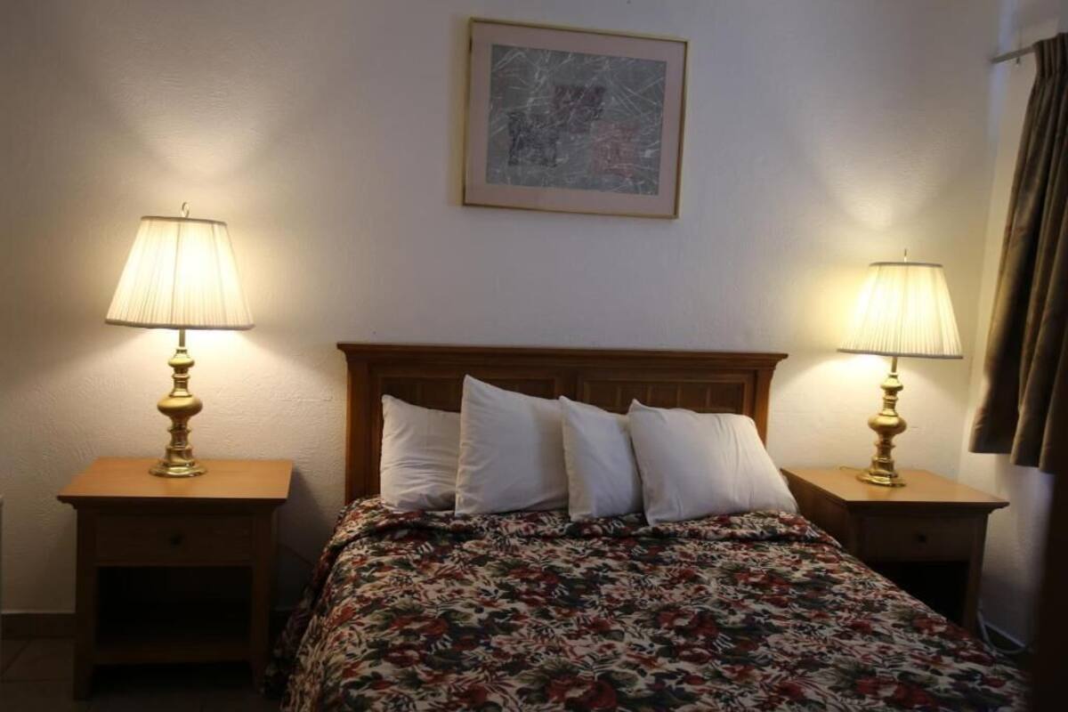 A neatly made bed with a floral bedspread, flanked by two bedside tables with lamps, in a room with plain walls and a single framed picture above the bed.