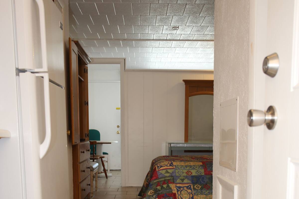 A narrow hallway with a tiled floor, a bed with a colorful quilt, a small desk, and a refrigerator.
