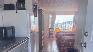 A narrow hallway with a kitchen area on one side, a bed with a red bedspread, and a view of buildings through a window. - El Ray motel (Wildwood)